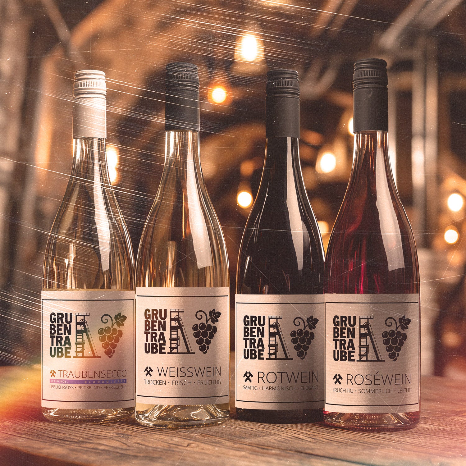 Vier Grubentraube-Flaschen nebeneinander im warm beleuchteten Keller: Traubensecco, Weißwein, Rotwein und Roséwein mit Etikett aus dem Ruhrpott (Zechenturm und Traube) – leblich-prickelnd, trocken-frisch, samtig-harmonisch und fruchtig-sommerlich.